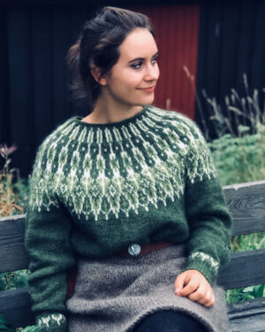 Jente som sitter på en benk ikledd en olivengrønn og grå strikkegenser av typen villmarksgenser