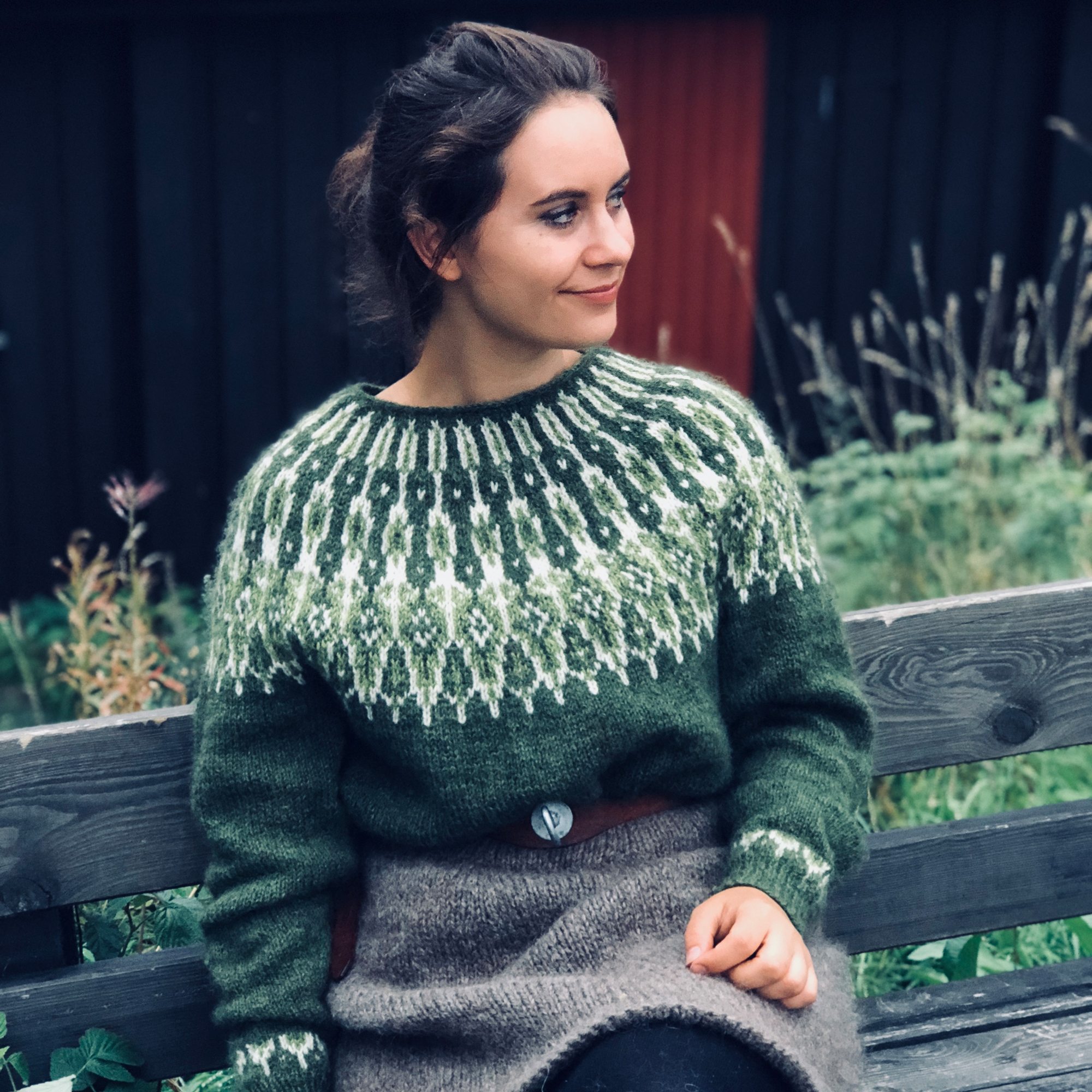 Jente som sitter på en benk ikledd en olivengrønn og grå strikkegenser av typen villmarksgenser